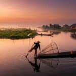 Un pêcheur en bateau traditionnel sur le lac Inle au lever du soleil avec des maisons sur pilotis en arrière-plan.