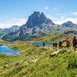 Un groupe de randonneurs marche sur un sentier de montagne avec le Pic du Midi d'Ossau en arrière-plan et des lacs bleus.